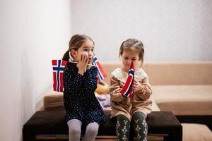 deux sœurs sont assises sur un canapé à la maison avec des drapeaux norvégiens sur les mains. norvège enfants filles avec drapeau. photo
