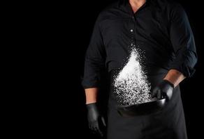 homme en uniforme noir tenant une poêle ronde en fonte avec du sel, le chef jette du sel blanc photo