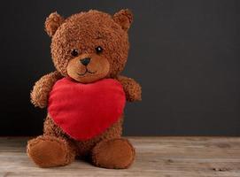 mignon ours en peluche brun tenant un gros coeur rouge et est assis sur un fond de bois noir photo