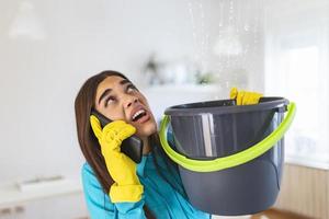 une jeune femme inquiète appelle un plombier pendant que l'eau de fuite tombe dans un seau à la maison photo