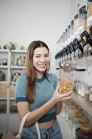 une cliente caucasienne remplit des aliments céréaliers dans un bocal en verre, lève le récipient réutilisable sur les étagères des magasins de recharge, une épicerie zéro déchet et sans plastique, des modes de vie durables et respectueux de l'environnement. photo