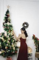 portrait d'une jolie jeune fille décorant un sapin de noël, souriante vêtue d'une robe rouge dans un salon de noël décoré à l'intérieur photo