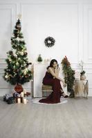 portrait de jolie jeune fille confortable s'asseoir, souriant porter une robe rouge dans le salon de noël décoré à l'intérieur photo