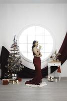 portrait d'une jolie jeune fille vêtue d'une robe rouge et buvant du vin, souriant à la caméra, debout dans un salon de noël décoré à l'intérieur photo