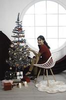 portrait de jolie jeune fille confortable s'asseoir, souriant porter une robe rouge dans le salon de noël décoré à l'intérieur photo