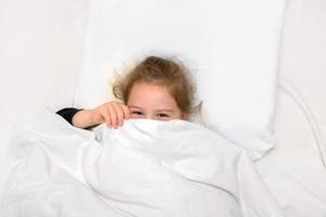 la petite fille au lit est recouverte d'une couverture, seuls les yeux rieurs sont visibles photo