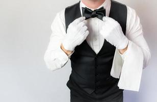 portrait de serveur ou majordome en gants blancs redressant le noeud papillon sur fond blanc. concept d'industrie de service et de courtoisie professionnelle. photo