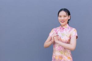 belle femme asiatique aux cheveux longs qui porte une robe cheongsam rose sur le thème du nouvel an chinois alors qu'elle montre sa main comme salutation chinoise avec sur fond gris. photo