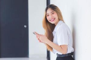 portrait d'une jolie étudiante thaïlandaise asiatique en uniforme sourit joyeusement tout en utilisant un smartphone à l'université. photo