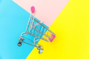 petit chariot d'épicerie de supermarché pour faire du shopping jouet avec roues isolé sur bleu jaune rose pastel coloré fond géométrique tendance espace de copie. vente acheter concept de consommateur de magasin de marché de centre commercial. photo