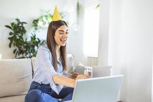 fête virtuelle. femme heureuse en chapeau de fête célébrant son anniversaire en ligne en quarantaine ou en auto-isolement, utilisant un ordinateur portable pour un appel vidéo avec des amis et la famille, tenant du champagne photo
