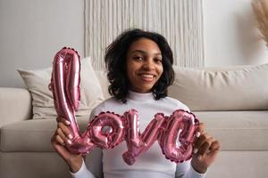 une fille souriante heureuse tient une inscription de ballon d'amour tout en étant assise près du canapé. appel vidéo photo