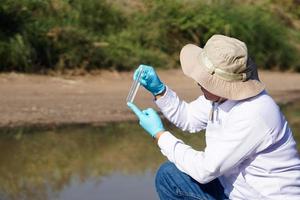 un chercheur en environnement d'homme asiatique tient un tube d'échantillon d'eau à inspecter au lac. concept, exploration, analyse de la qualité de l'eau de source naturelle. recherche de terrain en écologie. photo