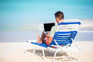 jeune homme avec ordinateur portable sur la plage tropicale. homme assis sur la chaise longue avec ordinateur photo