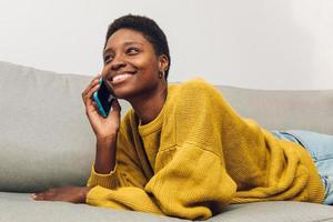 femme africaine utilisant un téléphone portable à la maison assise sur le canapé photo