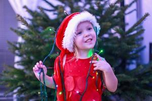 petite belle fille en robe rouge et chapeau avec des guirlandes de noël autour du cou photo