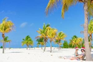 jeune maman et petite fille s'amusent sur une plage tropicale en bonnet de noel assis près d'un palmier photo