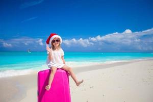 petite fille en bonnet de Noel assis sur une grande valise à la plage tropicale photo