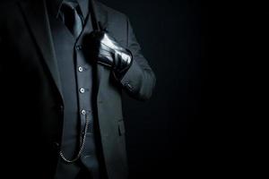 portrait d'un homme mystérieux en costume sombre et gants en cuir se tenant fièrement sur fond noir. concept de mafia hitman ou gentleman assassin. photo