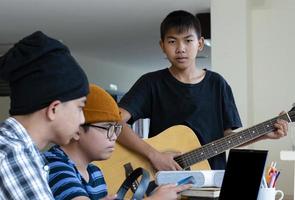 groupe de jeunes adolescents asiatiques assis ensemble à l'intérieur de la pièce travaillant et pratiquant un projet scolaire sur le sujet de la musique via un ordinateur portable et jouant du quitar, aussi, une mise au point douce et sélective. photo