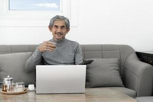 un homme âgé aux cheveux gris asiatiques en chemise à manches longues est assis sur un canapé et boit du thé chaud dans le salon de sa maison, concept pour une vie heureuse des aînés et une assurance-vie dans le monde entier. photo
