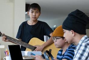 groupe de jeunes adolescents asiatiques assis ensemble à l'intérieur de la pièce travaillant et pratiquant un projet scolaire sur le sujet de la musique via un ordinateur portable et jouant du quitar, aussi, une mise au point douce et sélective. photo