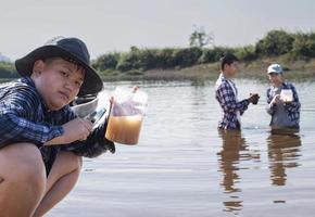 un jeune garçon asiatique tient un tube transparent qui a par exemple de l'eau à l'intérieur pour faire l'expérience et la mesure du niveau de ph pendant que son projet scolaire travaille avec ses amis derrière à la rivière où il vivait.