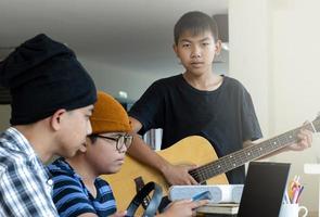 groupe de jeunes adolescents asiatiques assis ensemble à l'intérieur de la pièce travaillant et pratiquant un projet scolaire sur le sujet de la musique via un ordinateur portable et jouant du quitar, aussi, une mise au point douce et sélective. photo