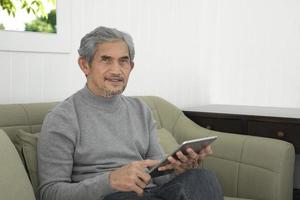 Portrait d'un homme asiatique senior âgé assis près d'une fenêtre en verre le matin pour travailler à domicile et vérifier ses affaires sur son ordinateur portable sur la table avec une mise au point sérieuse, douce et sélective. photo