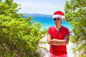 jeune homme au chapeau de noël rouge sur une plage tropicale près des sapins photo