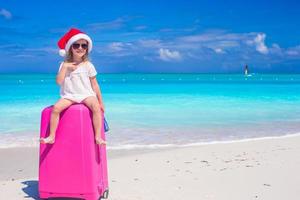 petite fille mignonne en bonnet de noel sur valise à la plage tropicale photo