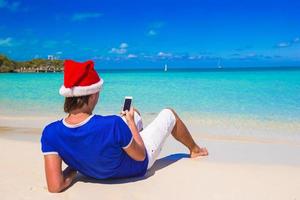 jeune homme en bonnet de noel parlant au téléphone sur une plage tropicale photo