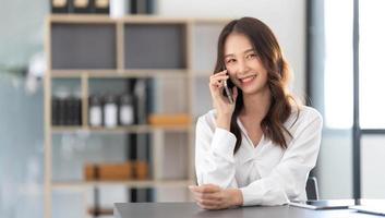 une jeune femme d'affaires asiatique s'assoit avec son ordinateur portable et parle joyeusement au téléphone avec son client expliquant les détails au bureau. photo
