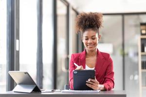 une jeune femme d'affaires afro-américaine assistante financière utilisant une tablette numérique et un ordinateur portable tout en travaillant sur un rapport financier.