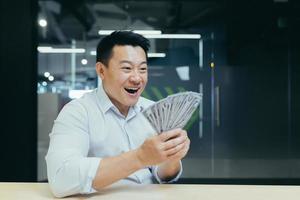 heureux jeune homme asiatique a reçu un salaire en espèces. il tient de l'argent dans ses mains, le montre, se réjouit photo