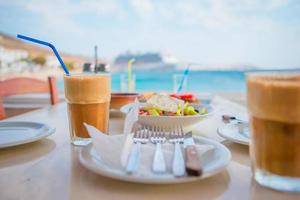 frappe sur le fond de table mer et grand coucheur. Frappuccino au café glacé d'été dans un grand verre et salade grecque au bar de la plage photo
