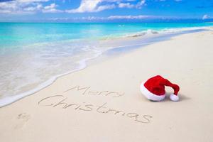 Joyeux Noël écrit sur le sable blanc de la plage avec un bonnet rouge photo