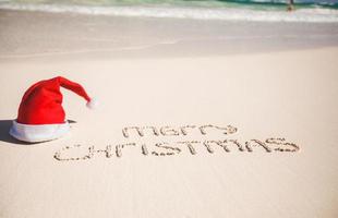 chapeau de père noël sur la plage de sable blanc et joyeux noël écrit dans le sable photo