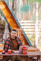 heureux jeune homme travaillant sur un ordinateur portable et buvant du café assis à la table en bois à l'extérieur photo