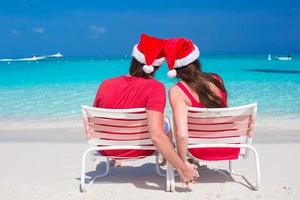 vue arrière du jeune couple en chapeaux de père noël rouges assis sur des chaises de plage photo