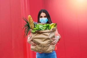 une jeune femme portant un masque médical tient un sac en papier écologique avec de la nourriture, des fruits et légumes, du poivre, de la baguette, de la laitue, une livraison intelligente en ligne sécurisée, un coronovirus, une quarantaine, une pandémie, un concept de séjour à la maison photo