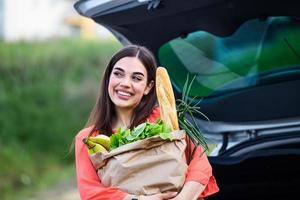 femme mettant ses sacs de shopping dans la voiture au parking du centre commercial. elle a fait quelques achats légers, principalement de la nourriture. jeune belle femme heureuse déplace l'achat du panier photo