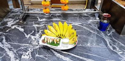 banane fraîche, petit verre d'eau et noix de bétel dans un plat blanc sur une table en marbre noir pour le culte statue de bouddha avec guirlande jaune accrochée à un cintre en acier inoxydable. offrandes, foi et concept alimentaire photo