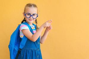 souriante petite écolière à lunettes, uniforme scolaire, sac à dos est attirée par l'attention pointant du doigt l'espace de copie. retour à l'école. publicité fournitures scolaires et papeterie photo