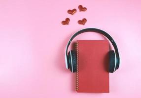 mise à plat d'un cahier rouge recouvert d'écouteurs sur fond rose décoré de coeurs scintillants rouges, espace de copie. livre audio, podcast, journal .love, saint valentin. photo