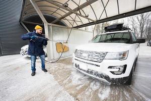 homme lavant une voiture suv américaine à eau haute pression avec galerie de toit au lavage en libre-service par temps froid. photo