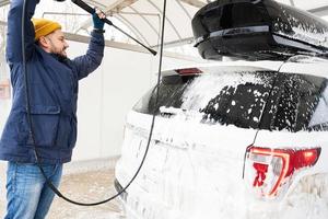 homme lavant une voiture suv américaine à eau haute pression avec galerie de toit au lavage en libre-service par temps froid. photo