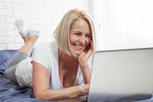 jolie femme souriante a un sourire agréable, une coupe de cheveux, vêtue de vêtements domestiques décontractés, rit en regardant une comédie, laisse des commentaires, discute en ligne avec des amis sur un ordinateur portable assis dans son lit photo