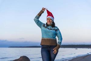 Heureuse belle jeune femme dans un chandail turquoise de chapeau de noël et un jean célébrant noël sur la plage photo