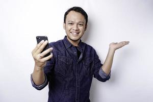 homme asiatique excité portant une chemise bleue pointant vers l'espace de copie à côté de lui tout en tenant son téléphone, isolé sur fond blanc photo